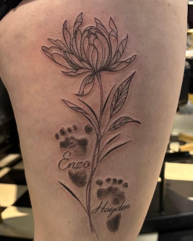 Fleur fineline avec deux empreintes de pieds de bébé et prénoms, sur la cuisse
